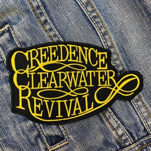 以下が含まれることがあります： 「Creedence Clearwater Revival」と黄色の刺繍が施された黒いパッチ。 パッチは装飾的なボーダーがあり、青いデニムの背景に配置されています。