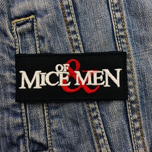 Könnte beinhalten: Ein rechteckiger schwarzer Aufnäher mit dem Text "OF MICE & MEN" in weißer und roter Stickerei. Der Aufnäher ist auf einer Jeansjacke befestigt, mit sichtbaren Nähten und einem Taschen Detail.