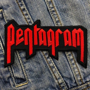 May include: A black embroidered patch with the word "Pentagram" in red, gothic-style lettering. The patch is rectangular with a unique, angular border and is set against a blue denim background.