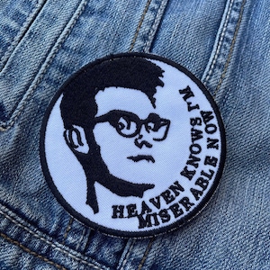 Op de afbeelding: Een ronde, witte patch met een zwarte illustratie van een persoon met een bril. De patch heeft de tekst "HEAVEN MISERABLE NOW I'M KNOWS" in een cirkelvormig patroon. De patch is op een blauw spijkerjasje.