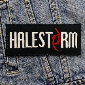 Könnte beinhalten: Ein rechteckiger schwarzer Aufnäher mit dem weißen gestickten Schriftzug "HALESTORM". Ein rotes stilisiertes Design befindet sich rechts vom Text. Der Aufnäher liegt auf einem blauen Jeansstoff.