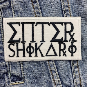 Puede incluir: Un parche rectangular blanco con letras y símbolos griegos bordados en negro. El parche está cosido a una chaqueta vaquera. El texto incluye las letras Σ, Π, T, Σ, R, Φ, K, Δ y Φ.
