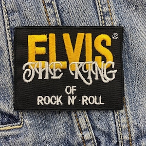 Op de afbeelding: Zwart rechthoekig embleem met gele geborduurde tekst "ELVIS" boven "THE KING OF ROCK 'N' ROLL" in wit. De patch is op een spijkerjas genaaid.