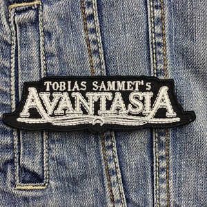 May include: A black and white embroidered patch with the text "TOBIAS SAMMET'S AVANTASIA" on a denim background. The patch has a black border and white lettering, and is rectangular in shape.