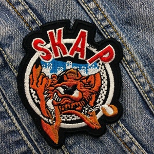 Op de afbeelding: Geborduurde patch met het woord "SKA" in rode letters. De patch toont een oranje en witte cartoon tijger met een skyline op de achtergrond. De patch is zwart en wit omrand.