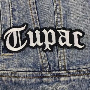 Puede incluir: Parche bordado con el nombre "Tupac" en letras góticas blancas sobre fondo negro. El parche está cosido sobre una chaqueta vaquera azul, un estilo clásico.
