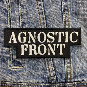 Puede incluir: Un parche rectangular negro con las palabras "AGNOSTIC FRONT" bordadas en blanco, sobre un fondo de mezclilla azul. El parche está diseñado para ser cosido en ropa, como una chaqueta o chaleco.