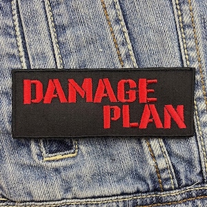 May include: A black rectangular patch featuring the red embroidered text "DAMAGE PLAN". The patch is attached to a blue denim jacket, with visible stitching and seams.