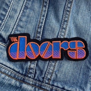 Könnte beinhalten: Gestickter Aufnäher mit dem Bandnamen "The Doors" in Orange und Blau. Der Aufnäher ist schwarz umrandet und auf eine Jeansjacke genäht. Der Text hat einen Farbverlauf, der von Blau nach Lila übergeht.