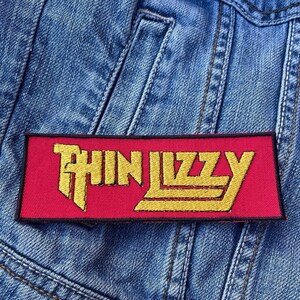 Può includere: Toppa rettangolare con la scritta "THIN LIZZY" in oro su sfondo rosso. La toppa è bordata di nero ed è applicata su una giacca di jeans blu.