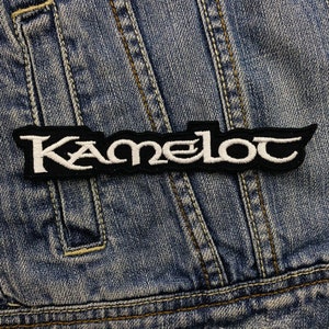 Könnte beinhalten: Ein schwarz-weißer, gestickter Aufnäher mit dem Wort "Kamelot" in einer stilisierten Schriftart. Der Aufnäher ist rechteckig mit gezackten Kanten und befindet sich auf einer Jeansjacke.
