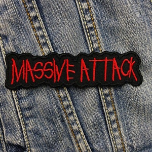 Könnte beinhalten: Ein schwarzer, gestickter Aufnäher mit den roten Worten "MASSIVE ATTACK", auf eine Jeansjacke genäht. Der Aufnäher hat einen wellenförmigen Rand.