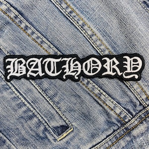 Könnte beinhalten: Ein schwarz-weißer, gestickter Aufnäher mit dem Wort "BATHORY" in gotischer Schrift. Der Aufnäher ist rechteckig und hat einen weißen Rand. Er befindet sich auf einer blauen Jeansjacke.