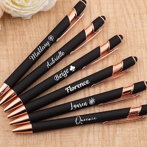 Puede incluir: Un conjunto de bolígrafos negros con detalles en oro rosa, cada uno grabado con un nombre en escritura blanca. Los bolígrafos están dispuestos sobre una superficie de madera clara, con flores en el fondo.