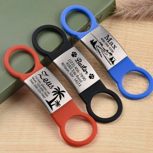 Op de afbeelding: Drie gepersonaliseerde hondenpenningen in rood, zwart en blauw. Elke penning heeft een metalen plaat met gegraveerde tekst en een rubberen lus. De rode penning zegt "Zeus" met een palmboomafbeelding. De zwarte penning zegt "Buster" met pootafdrukken.