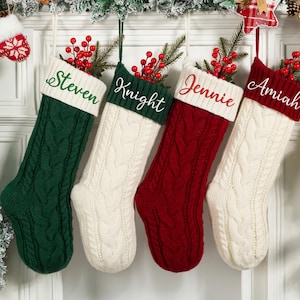 May include: Four cable-knit Christmas stockings in green, cream, and red, each with a white cuff embroidered with a name: Steven, Knight, Jennie, and Amiah. The stockings are decorated with sprigs of red berries and greenery.