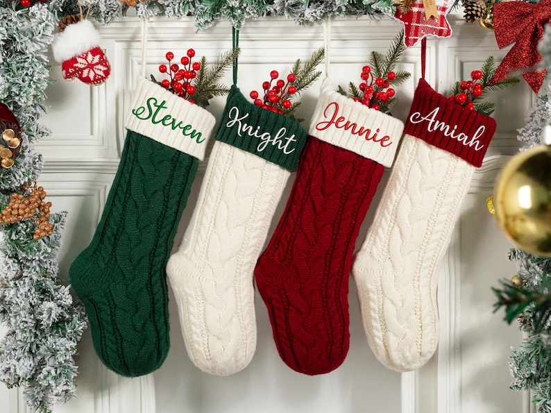 May include: Four cable-knit Christmas stockings in green, cream, and red, each with a white cuff embroidered with a name: Steven, Knight, Jennie, and Amiah. The stockings are decorated with sprigs of red berries and greenery.