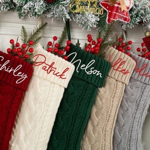 May include: A collection of six cable-knit Christmas stockings in various colors, including red, cream, green, tan, and gray. Each stocking is personalized with a name embroidered in a contrasting color. The stockings are decorated with red berries and greenery.