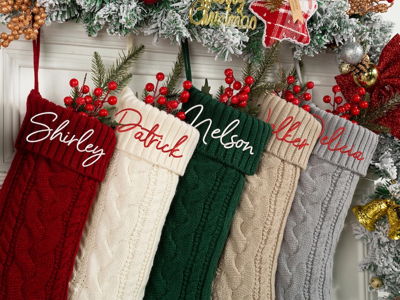 May include: A collection of six cable-knit Christmas stockings in various colors, including red, cream, green, tan, and gray. Each stocking is personalized with a name embroidered in a contrasting color. The stockings are decorated with red berries and greenery.