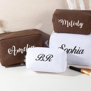 May include: A selection of personalised cosmetic bags in brown and white. The brown bags have white cursive text that reads "Amelia" and "Melody." The white bags have black text that reads "Sophia" and "B.R." Makeup brushes are visible.