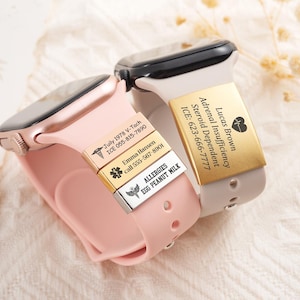 May include: Two silicone smartwatch bands in light pink and beige, each featuring custom metal medical alert tags. The pink band has three stacked silver and gold tags with engraved text including names, phone numbers, and allergy information.