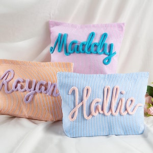 May include: Three rectangular pouches made of striped seersucker fabric. One orange and white pouch features the name Rayan in purple yarn. One pink and white pouch features the name Maddy in blue yarn. One blue and white pouch features the name Hallie in pink yarn.