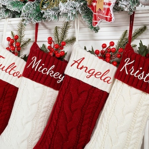 May include: Four cable-knit Christmas stockings in red and white hang from a decorated mantel. Each stocking has a white cuff with a name embroidered in red: Fulton, Mickey, Angela, and Kristin. Red berries and greenery adorn the top.