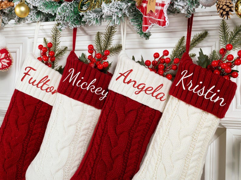 May include: Four cable-knit Christmas stockings in red and white hang from a decorated mantel. Each stocking has a white cuff with a name embroidered in red: Fulton, Mickey, Angela, and Kristin. Red berries and greenery adorn the top.