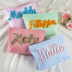 May include: Four rectangular zipped pouches in pink, green, red, and blue pinstriped fabric. Each pouch features a raised, cursive name in a contrasting colour: Maddy in blue, Fillippa in orange, Kate in green, and Hallie in light pink.