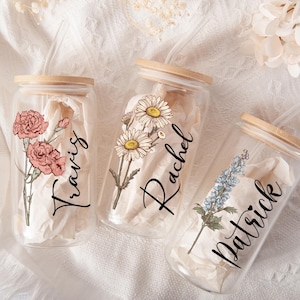 May include: Three glass beverage jars with wooden lids, each adorned with floral illustrations and names: "Travis," "Rachel," and "Patrick." The jars are filled with white paper and sit on a white, textured surface.