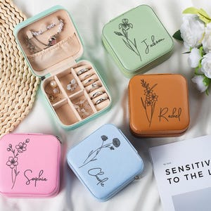 Pode incluir: Uma coleção de pequenas caixas de joias quadradas em várias cores: rosa, azul claro, verde e castanho. Cada caixa apresenta um design floral e um nome. Uma caixa está aberta, revelando joias. As caixas estão sobre uma superfície branca.