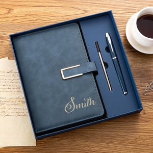 Peut inclure: Un carnet bleu foncé avec une fermeture métallique dorée et le nom Smith gravé sur la couverture, présenté dans un coffret cadeau bleu assorti avec un stylo à bille bleu foncé et une recharge d'encre argentée, posé sur une surface en bois.