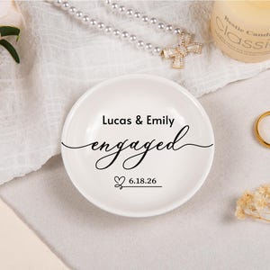 Pode incluir: Um prato redondo de cer&acirc;mica branca com texto impresso em preto que diz Lucas & Emily engaged e a data 18.06.26 com um pequeno &iacute;cone de cora&ccedil;&atilde;o. O prato tem uma superf&iacute;cie lisa e uma forma circular simples para guardar pequenos objetos.