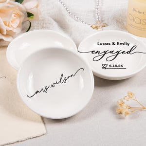 Pode incluir: Dois pratos de cer&acirc;mica branca para an&eacute;is com texto em preto. Um prato apresenta o texto "mrs.wilson" em letra cursiva. O outro exibe o texto "Lucas & Emily", a palavra "engaged" em cursivo, um &iacute;cone de cora&ccedil;&atilde;o e a data "6.18.26" em preto.