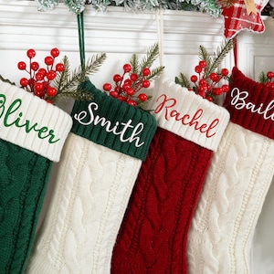 Könnte beinhalten: Vier Zopfstrick-Weihnachtsstrümpfe in Grün, Rot und Weiß, jeder mit einem Namen auf der Manschette: Oliver, Smith, Rachel und Bailey. Die Strümpfe sind mit roten Beeren und Grünzeug verziert.