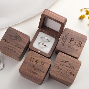 May include: A collection of square dark walnut wood ring boxes with hinged lids. Each box features custom laser-engraved names, dates, and graphics including rings, mountains, and foliage. One open box displays two silver rings on a white velvet insert.
