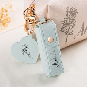 Può includere: Una trousse cosmetica rosa chiaro con il nome Anna e un'illustrazione floreale, abbinata a un astuccio per rossetto azzurro con il nome Natalie e un ciondolo a forma di cuore. Entrambi gli articoli presentano chiusure e dettagli in metallo dorato.