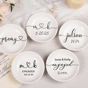 Pode incluir: Cinco pratos de cer&acirc;mica branca para an&eacute;is com texto e gr&aacute;ficos personalizados em preto. Os pratos exibem nomes como Jeremy, Julian, Lucas & Emily, iniciais m & k, e datas como 5.20.25, 02.14.21 e 6.18.26, decorados com motivos de cora&ccedil;&otilde;es.