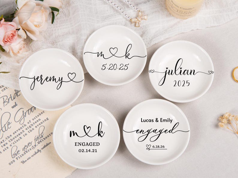 Pode incluir: Cinco pratos de cer&acirc;mica branca para an&eacute;is com texto e gr&aacute;ficos personalizados em preto. Os pratos exibem nomes como Jeremy, Julian, Lucas & Emily, iniciais m & k, e datas como 5.20.25, 02.14.21 e 6.18.26, decorados com motivos de cora&ccedil;&otilde;es.