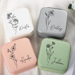 Könnte beinhalten: Vier quadratische Schmuckkästchen in Weiß, Grau, Rosa und Grün. Jedes Etui hat ein Blumendesign und einen Namen in Schreibschrift. Die Etuis sind mit Reißverschluss versehen und scheinen aus Kunstleder zu sein.