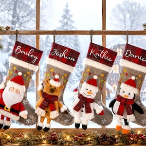 May include: Four grey felt Christmas stockings hanging in a row, each featuring a red and black plaid cuff with the names Bailey, Joshua, Kathie, and Daniel embroidered in white. Each stocking displays a plush character: Santa, reindeer, snowman, and penguin.