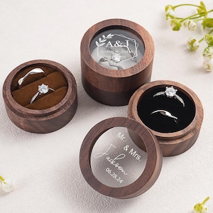 Puede incluir: Tres cajas redondas para anillos de madera de nogal oscuro con tapas. Una tapa presenta el texto grabado A&J Aiden & Julia, y otra Mr. & Mrs. Jackson 06.28.24. Las cajas contienen anillos de plata con piedras claras sobre terciopelo.