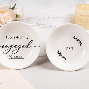 Pode incluir: Dois pratos de cer&acirc;mica branca para an&eacute;is. O prato esquerdo apresenta o texto Lucas & Emily, a palavra engaged em letra cursiva e a data 6.18.26 com um &iacute;cone de cora&ccedil;&atilde;o. O direito mostra as iniciais D T separadas por um cora&ccedil;&atilde;o e dois ramos.