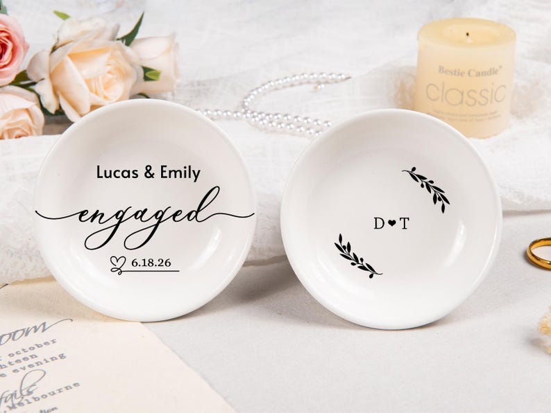 Pode incluir: Dois pratos de cer&acirc;mica branca para an&eacute;is. O prato esquerdo apresenta o texto Lucas & Emily, a palavra engaged em letra cursiva e a data 6.18.26 com um &iacute;cone de cora&ccedil;&atilde;o. O direito mostra as iniciais D T separadas por um cora&ccedil;&atilde;o e dois ramos.