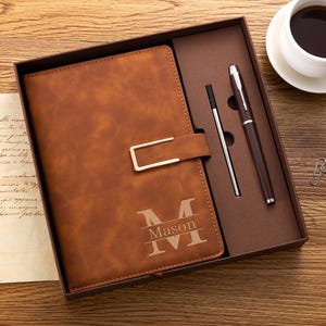 May include: A brown faux leather notebook with a gold-toned metal clasp and the engraved name Mason over a large letter M. The notebook is presented in a brown gift box alongside a matching brown ballpoint pen and a silver metal pen refill.