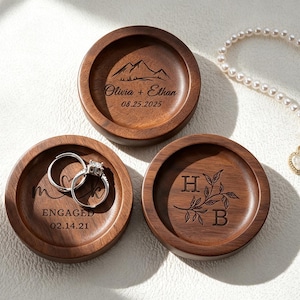 Pode incluir: Três pratos redondos de madeira castanha escura para anéis com gravações a laser personalizadas. Um apresenta um design de montanha com "Olivia + Ethan 08.25.2025", outro "ENGAGED 02.14.21" com dois anéis de prata e o terceiro as iniciais "H" e "B".