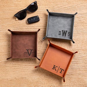 May include: Three square valet trays in grey, brown, and tan, each featuring a personalised monogram. The grey tray displays DWS, the brown tray shows a V, and the tan tray features KT. Black sunglasses and a car key fob are placed nearby.