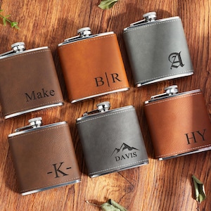 May include: Six personalised hip flasks in shades of brown and grey, arranged on a wooden surface. Each flask has a silver cap and is engraved with initials, names, or a mountain graphic with the name "Davis".