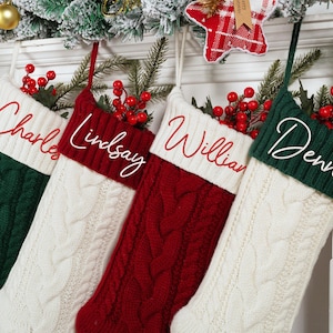 May include: Four cable-knit Christmas stockings in green, white, and red, each with a name embroidered in white script: Charles, Lindsay, William, and Dennis. The stockings are decorated with red berries and greenery.