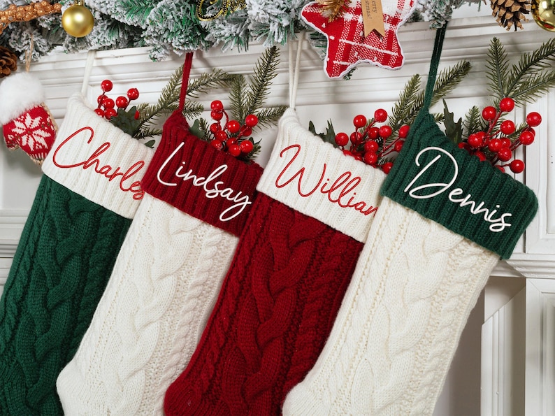 May include: Four cable-knit Christmas stockings in green, white, and red, each with a name embroidered in white script: Charles, Lindsay, William, and Dennis. The stockings are decorated with red berries and greenery.